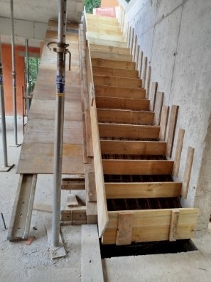 Szczegółowe ujęcie drewnianego szalunku schodów żelbetowych z widocznym zbrojeniem, wspartego na stemplach budowlanych, na tle surowej betonowej ściany.