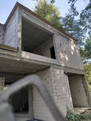 Dwukondygnacyjny budynek w trakcie budowy, ściany wzniesione z białych bloczków, widoczne zbrojenia, w tle zieleń drzew.