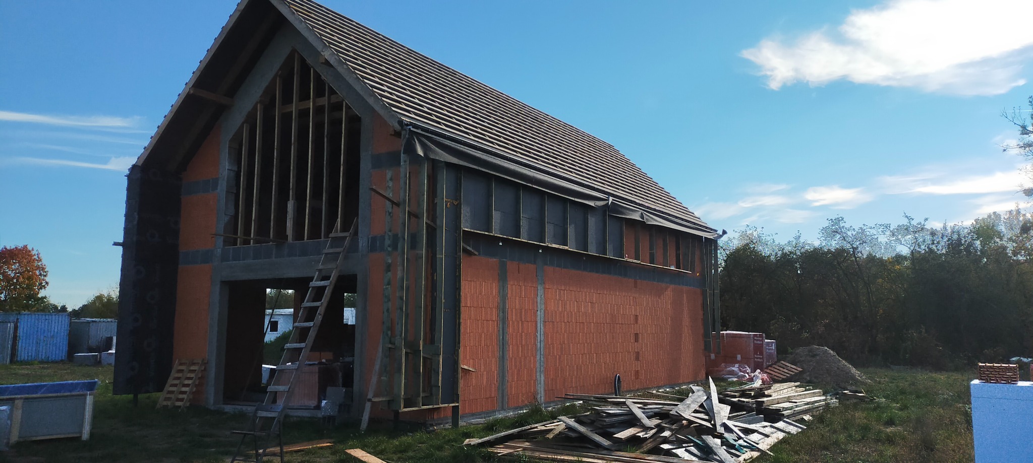 Budowa domu w stanie surowym otwartym: widoczne ściany z czerwonej cegły, drewniana konstrukcja dachu z pokryciem, drabina oparta o ścianę, materiały budowlane na trawie, błękitne niebo.