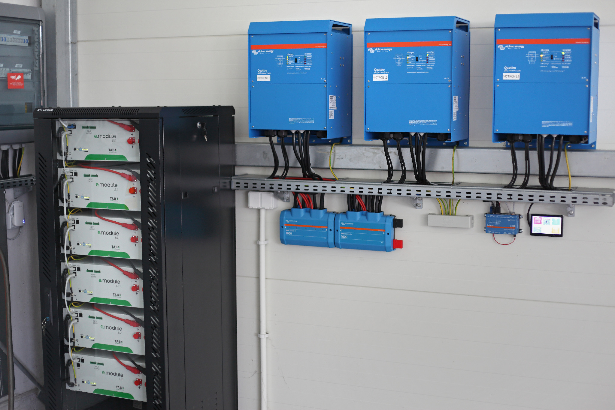 Instalacja magazynu energii: rząd niebieskich inwerterów Victron Energy Quattro L2 i L3 nad dwoma mniejszymi inwerterami, obok szafa z modułami bateryjnymi TAB e.module L5.1, widoczny panel...