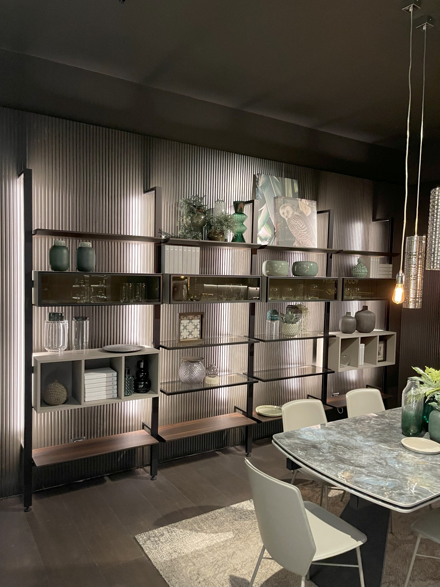 Regał modułowy z półkami szklanymi i drewnianymi, wypełniony dekoracjami, w nowoczesnym salonie w Gdyni, oświetlony wiszącymi lampami.