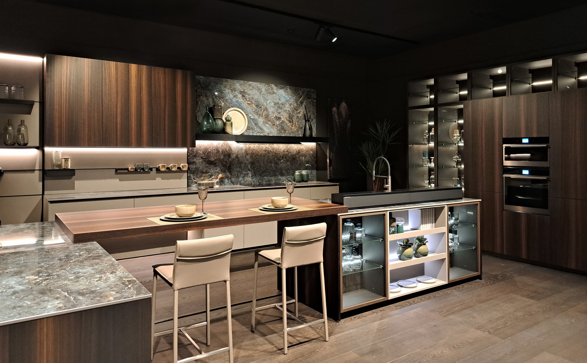 Nowoczesna kuchnia z wyspą, drewnianymi frontami szafek i blatem z kamienia, oświetleniem LED i barkiem z przeszklonymi półkami.