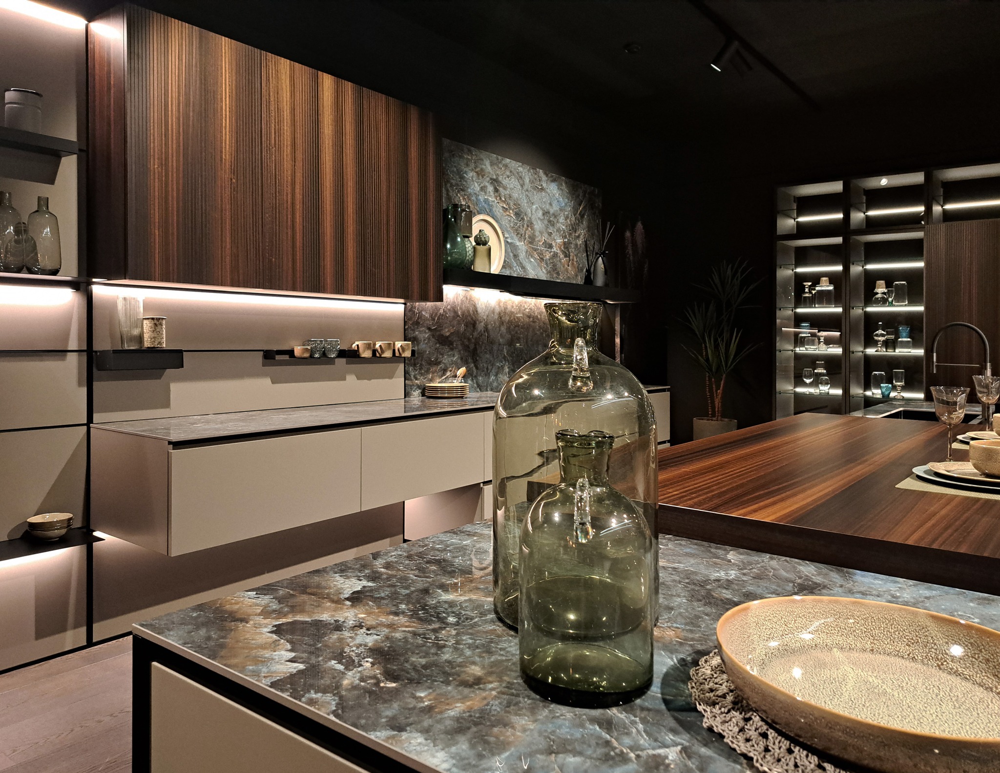 Nowoczesna kuchnia z drewnianymi frontami szafek i blatem z marmurowym wzorem, oświetlenie LED pod szafkami, szklane naczynia dekoracyjne na blacie.