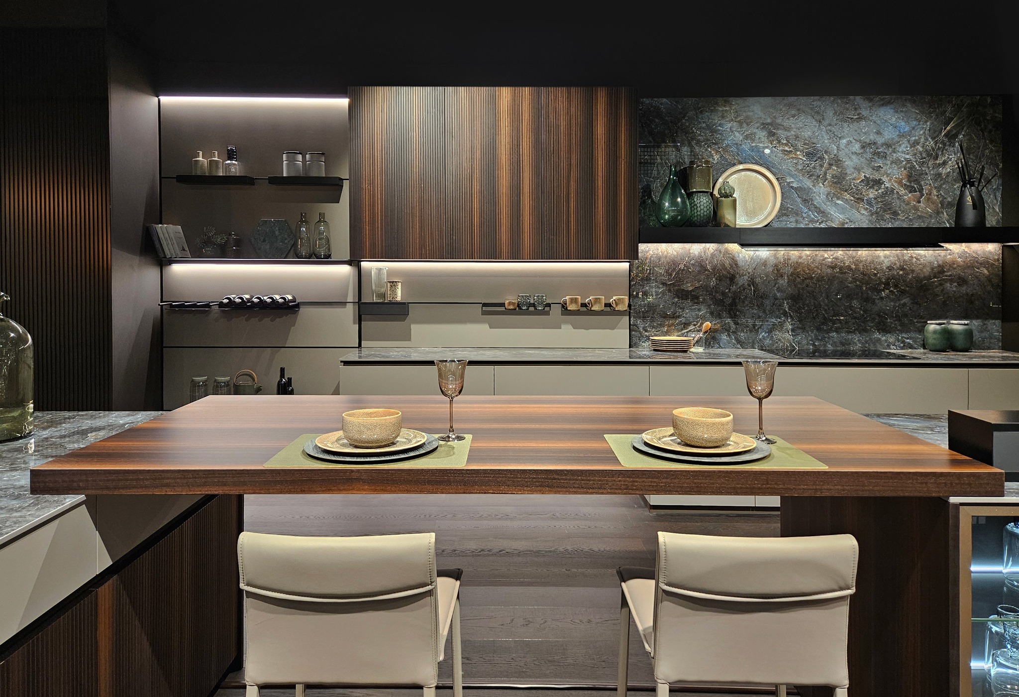 Nowoczesna kuchnia z wyspą pełniącą funkcję stołu jadalnego, nakrycia dla dwóch osób, fronty szafek z ciemnego drewna z wyraźnym usłojeniem, marmurowy backsplash, oświetlenie LED pod szafkami.