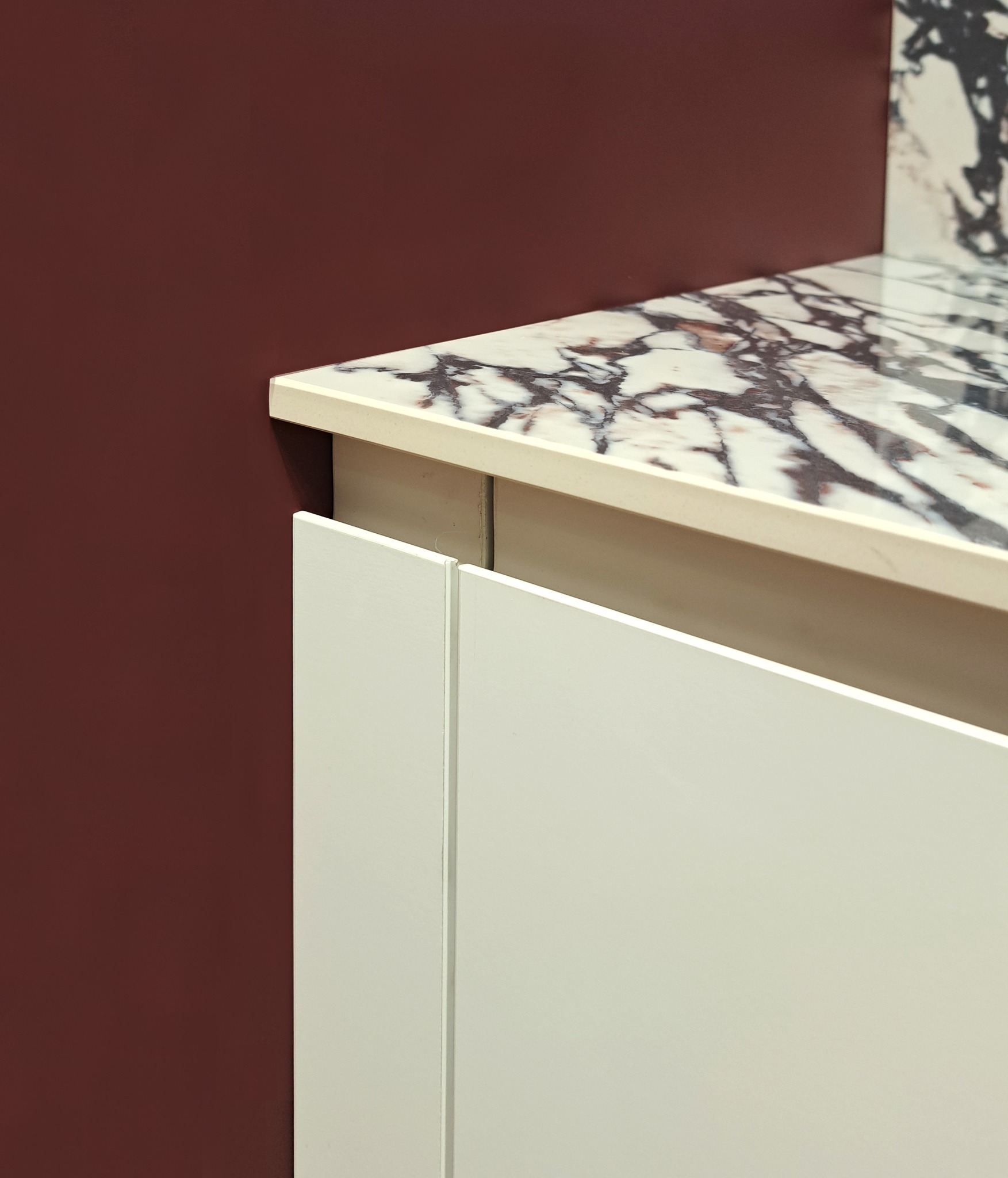 Fragment blatu kuchennego z marmurowym wzorem i białymi frontami szafek na tle bordowej ściany.