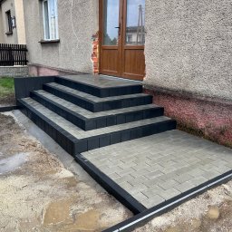 MAJZEL PROJEKT KRZYSZTOF MAJZEL - Nowe schody z kostki brukowej w odcieniach szarości i czerni prowadzące do drzwi wejściowych domu. Widoczne ślady prac ziemnych wokół.