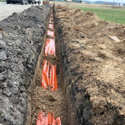 MAJZEL PROJEKT KRZYSZTOF MAJZEL - Wykopany wąski rów w ziemi z pomarańczowymi osłonami kabli, przygotowany pod instalację. Widoczna struktura gleby i krawędzie wykopu. Prace ziemne w toku.