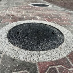 MAJZEL PROJEKT KRZYSZTOF MAJZEL - Ułożona kostka brukowa w odcieniach szarości i czerwieni, z okrągłymi włazami studzienek kanalizacyjnych obramowanymi jasnym, drobnym kruszywem.