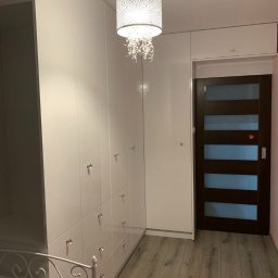 ARTESIA PROSTA SPÓŁKA AKCYJNA - Biała szafa na wymiar z szufladami i ozdobnymi uchwytami w mieszkaniu we Wrocławiu, oświetlona dekoracyjną lampą wiszącą, widoczna rama łóżka.