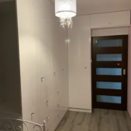 Biała szafa na wymiar z szufladami i ozdobnymi uchwytami w mieszkaniu we Wrocławiu, oświetlona dekoracyjną lampą wiszącą, widoczna rama ł&oacute;żka.