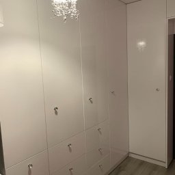 ARTESIA PROSTA SPÓŁKA AKCYJNA - Biała szafa wnękowa z połyskiem, srebrne gałki, drewniana podłoga i ozdobny żyrandol zwisający z sufitu.