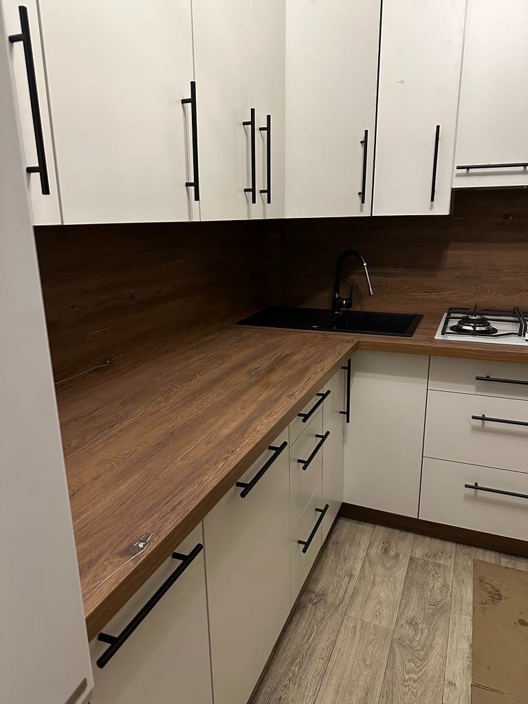 Nowoczesna kuchnia z białymi szafkami, drewnianym blatem i czarnymi uchwytami, widok na zlew i płytę gazową.