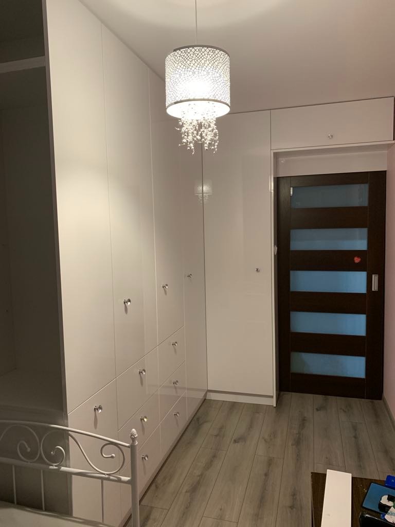 Biała szafa na wymiar z szufladami i ozdobnymi uchwytami w mieszkaniu we Wrocławiu, oświetlona dekoracyjną lampą wiszącą, widoczna rama łóżka.