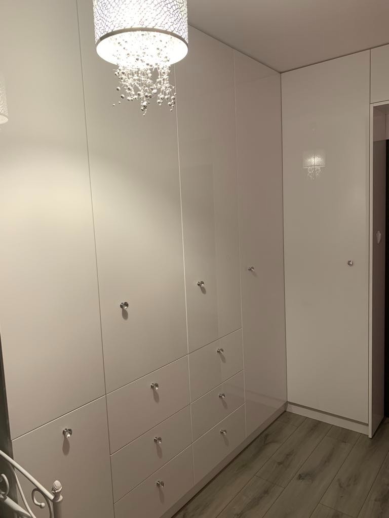 Biała szafa wnękowa z połyskiem, srebrne gałki, drewniana podłoga i ozdobny żyrandol zwisający z sufitu.