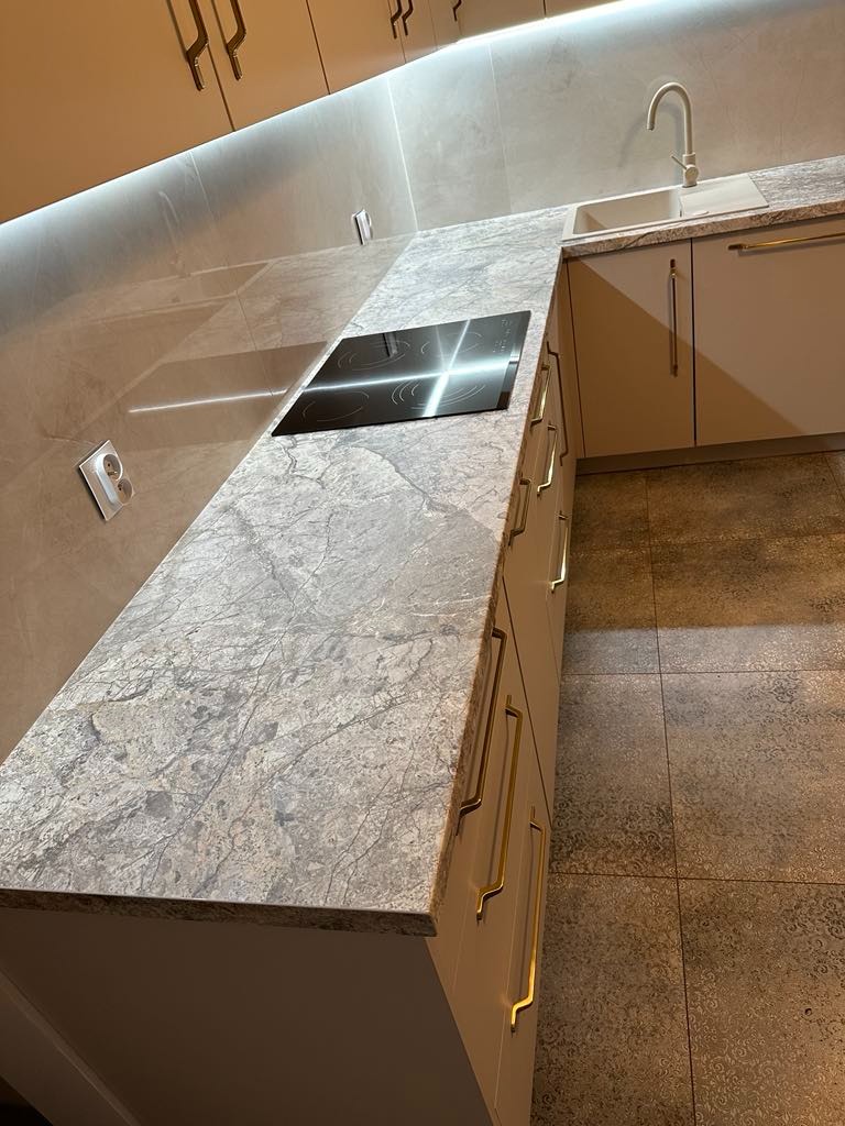 Jasna kuchnia z blatem z szarego marmuru, płyta indukcyjna i złote uchwyty szafek, oświetlenie LED podszafkowe.