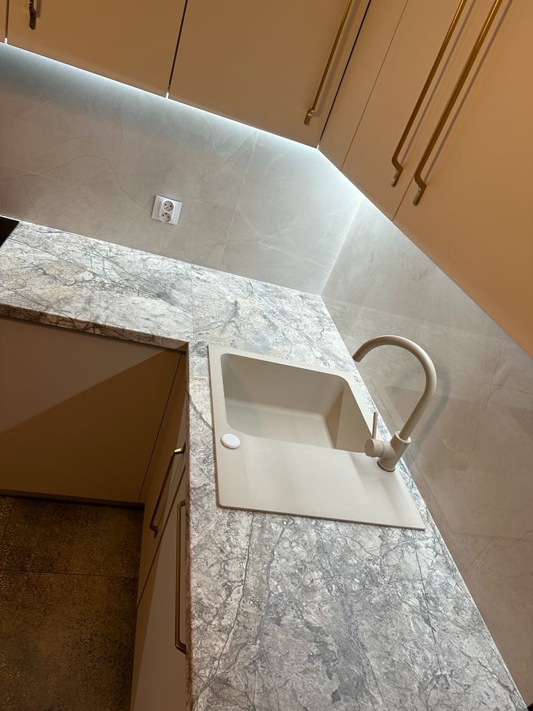 Beżowy zlew z baterią wkomponowany w blat o marmurowej fakturze, widok z góry, szafki kuchenne w tle z ozdobnymi uchwytami.