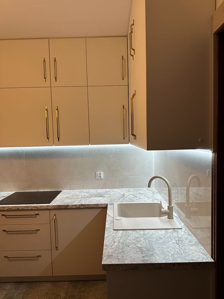 Jasna kuchnia z beżowymi szafkami z długimi złotymi uchwytami, marmurowy blat i zlew w tym samym kolorze, podświetlenie LED pod szafkami wiszącymi.