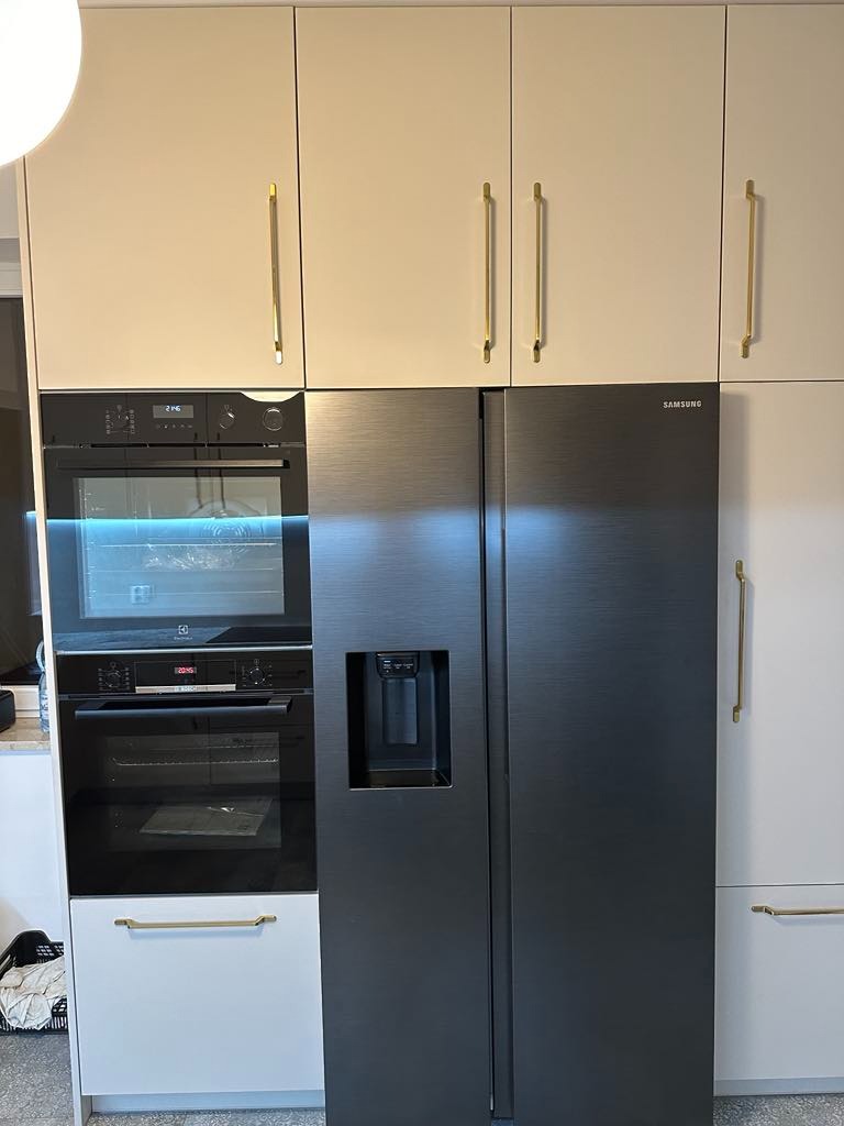 Nowoczesna zabudowa kuchenna z beżowymi szafkami górnymi, czarnym piekarnikiem i czarną lodówką Samsung z dyspenserem wody, uchwyty w kolorze złotym.