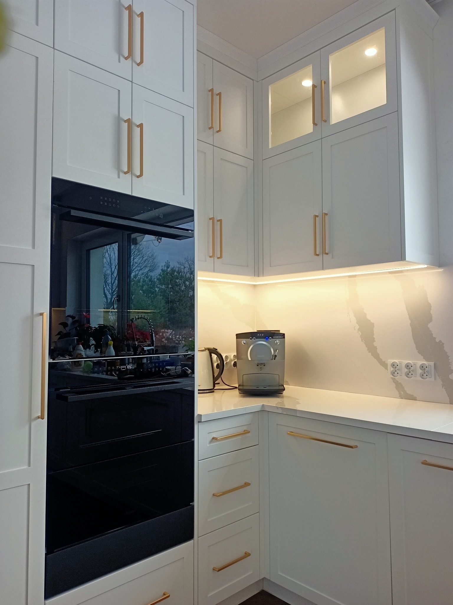 Biała kuchnia z zabudową, złote uchwyty, oświetlenie LED pod szafkami, ekspres do kawy na blacie. Widoczny piekarnik w zabudowie i okno odbijające się w szybie.