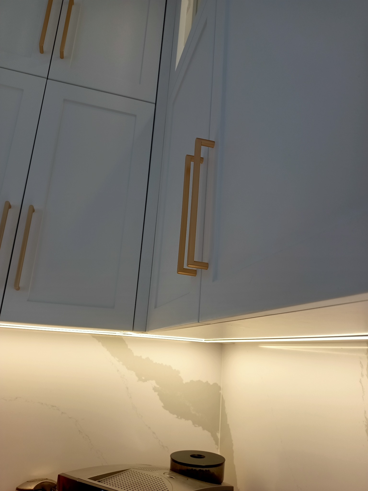 Białe szafki kuchenne z podświetleniem LED i złotymi uchwytami, widoczne z dołu, z blatem imitującym marmur i fragmentem ekspresu do kawy.