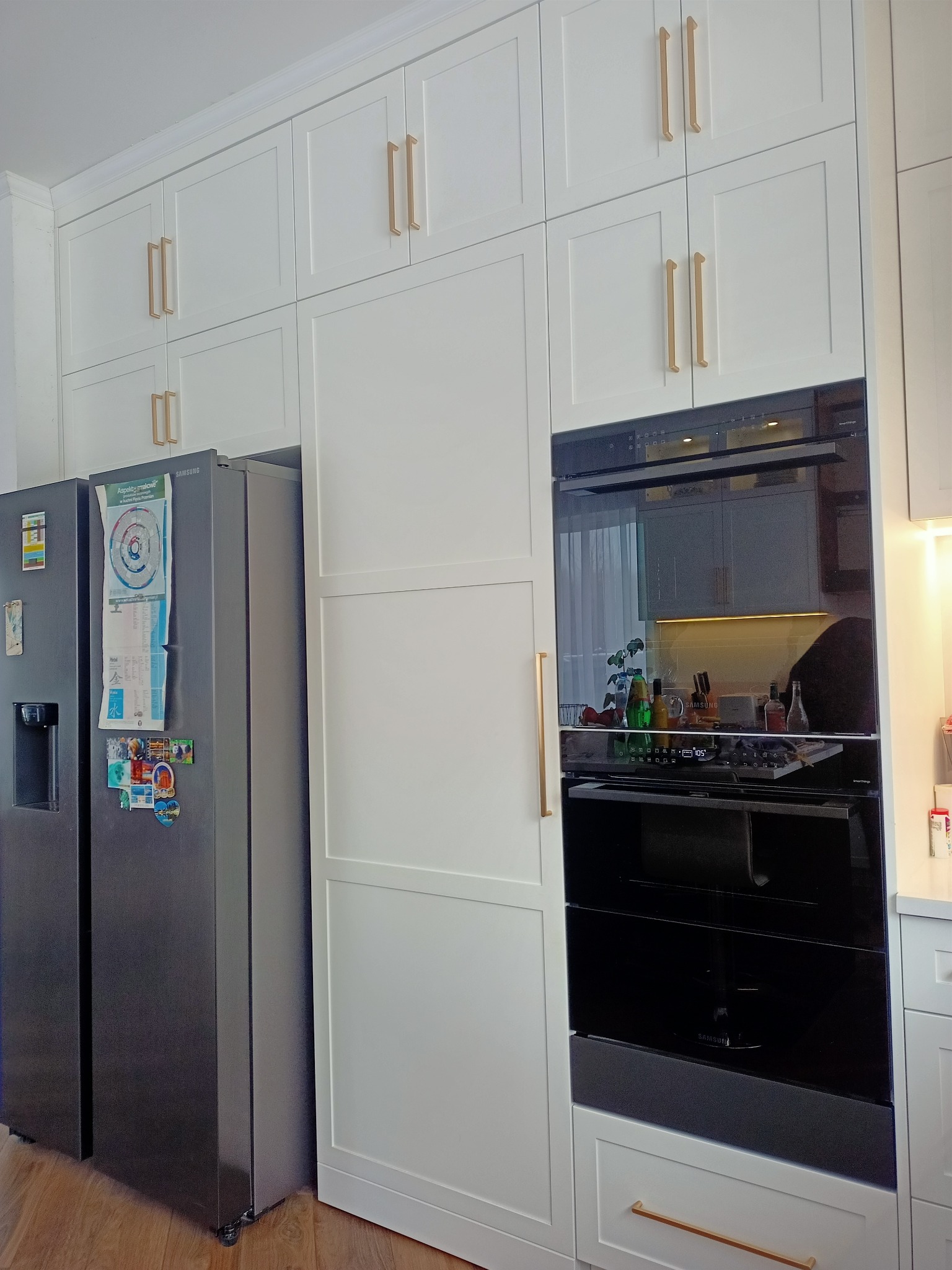 Biała zabudowa kuchenna z piekarnikiem i lodówką Samsung, złote uchwyty, minimalistyczny design, nowoczesne wykończenie, widok na wnętrze przez szybę piekarnika.