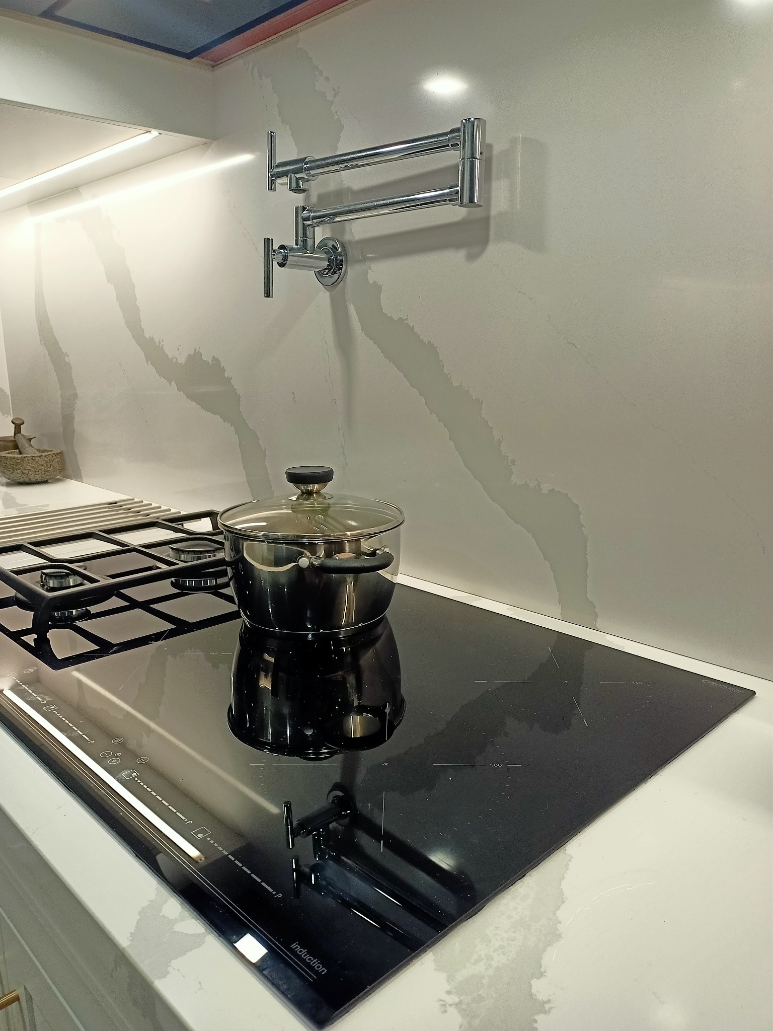 Nowoczesna kuchnia z czarną płytą indukcyjną, garnkiem pod szklaną pokrywką i baterią nad płytą na tle ściany z białego marmuru z szarymi żyłami.