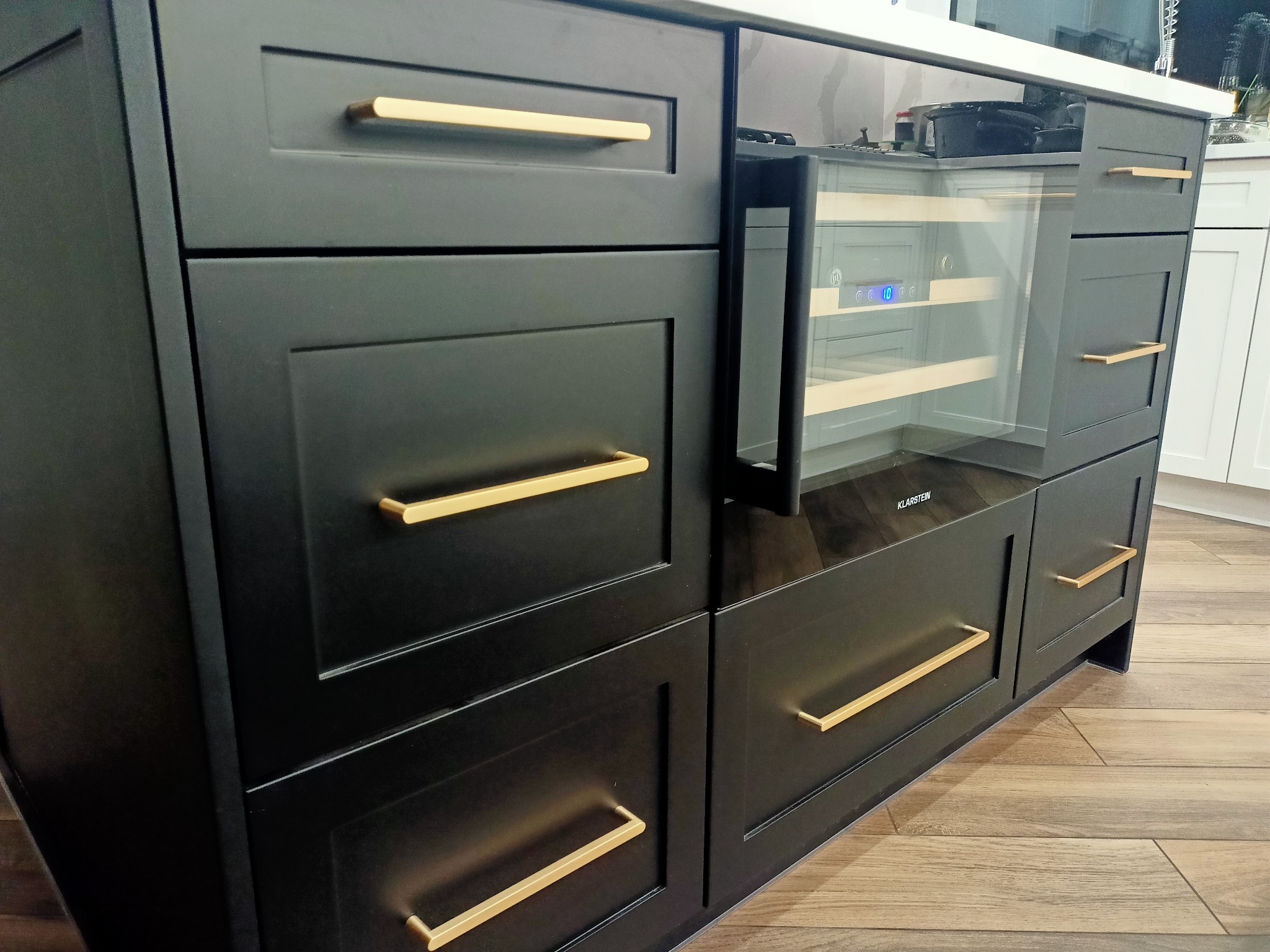 Czarna zabudowa kuchenna z złotymi uchwytami i wbudowaną chłodziarką do wina Klarstein, widoczna na tle drewnianej podłogi i fragmentu białej zabudowy.