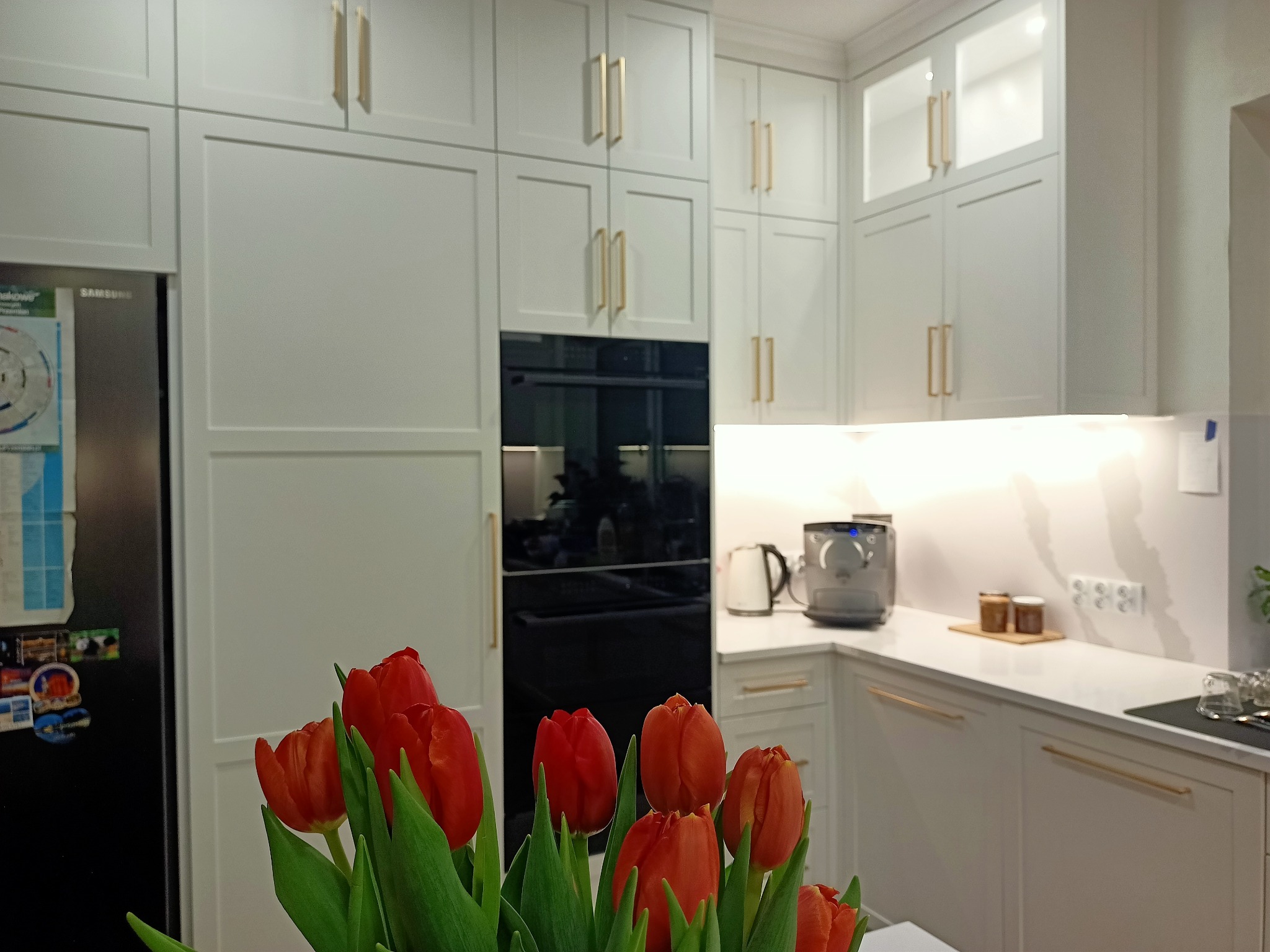 Jasna kuchnia z frezowanymi frontami i złotymi uchwytami, z czarnym piekarnikiem w zabudowie. Na pierwszym planie bukiet czerwonych tulipanów.