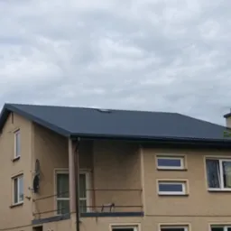 Dwupiętrowy dom z nowym, ciemnoszarym dachem z blachy trapezowej i oknem dachowym, na tle pochmurnego nieba. Widoczne rynny i komin.