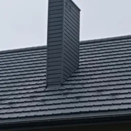 Szary, metalowy komin wentylacyjny na dachu pokrytym ciemną dachówką. Widoczne obróbki blacharskie wokół komina. Dachówka ceramiczna tworzy regularny wzór.