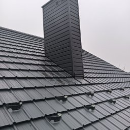 KRIS KRZYSZTOF HOBER - Szary, metalowy dach z kominem i zabezpieczeniami przeciwśniegowymi w pochmurny dzień. Widok z góry na nowo pokryty dach z blachy trapezowej.