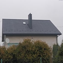 KRIS KRZYSZTOF HOBER - Dom z nowym, ciemnoszarym dachem z dachówki i kominem. Widoczne rynny, okno dachowe i antena satelitarna. Tło: pochmurne niebo, roślinność.