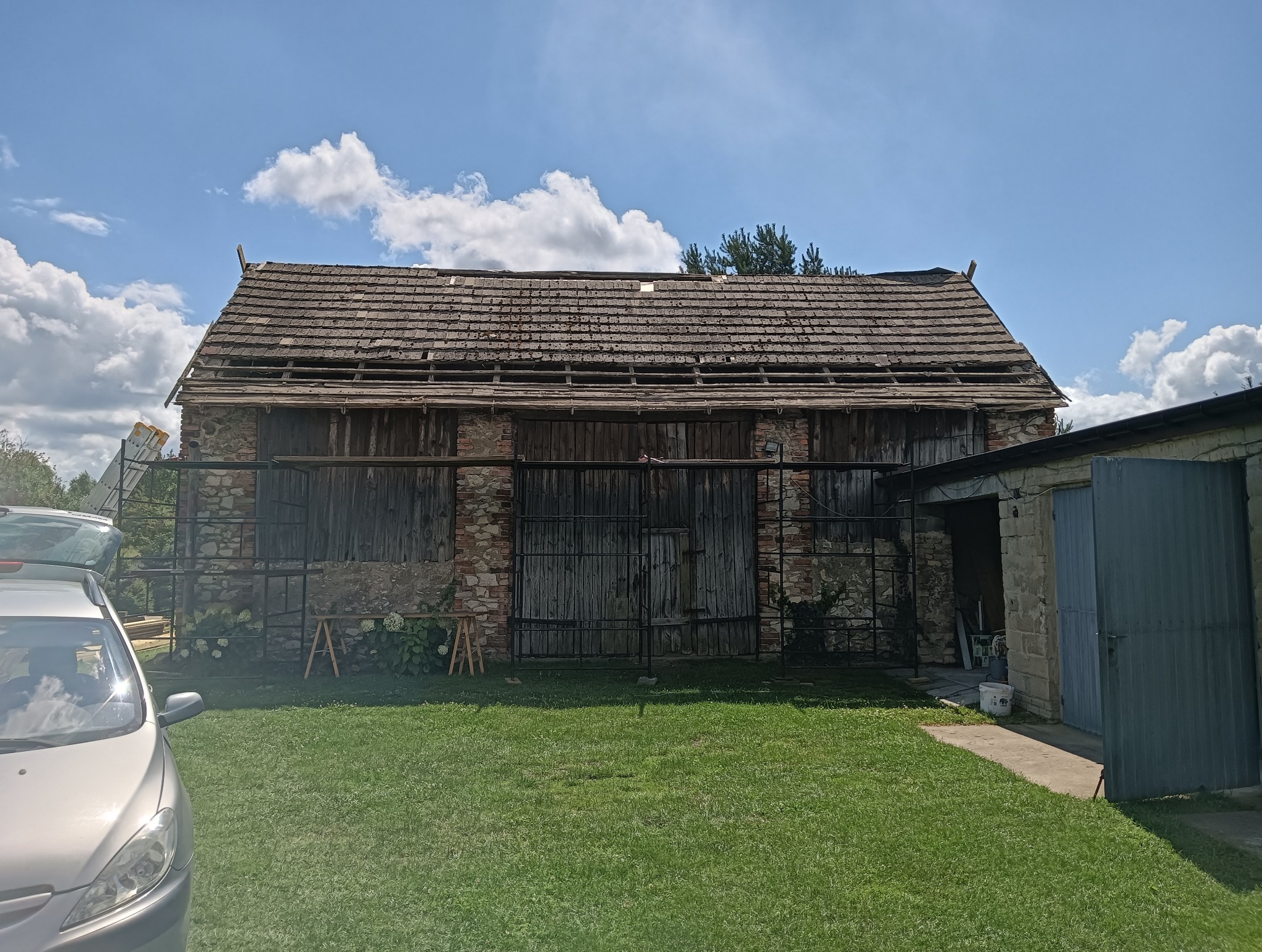 Stary, częściowo zniszczony dach z drewnianej dachówki na budynku z kamienia i drewna. Widoczne ubytki w pokryciu i elementy konstrukcji. W tle błękitne niebo z chmurami.