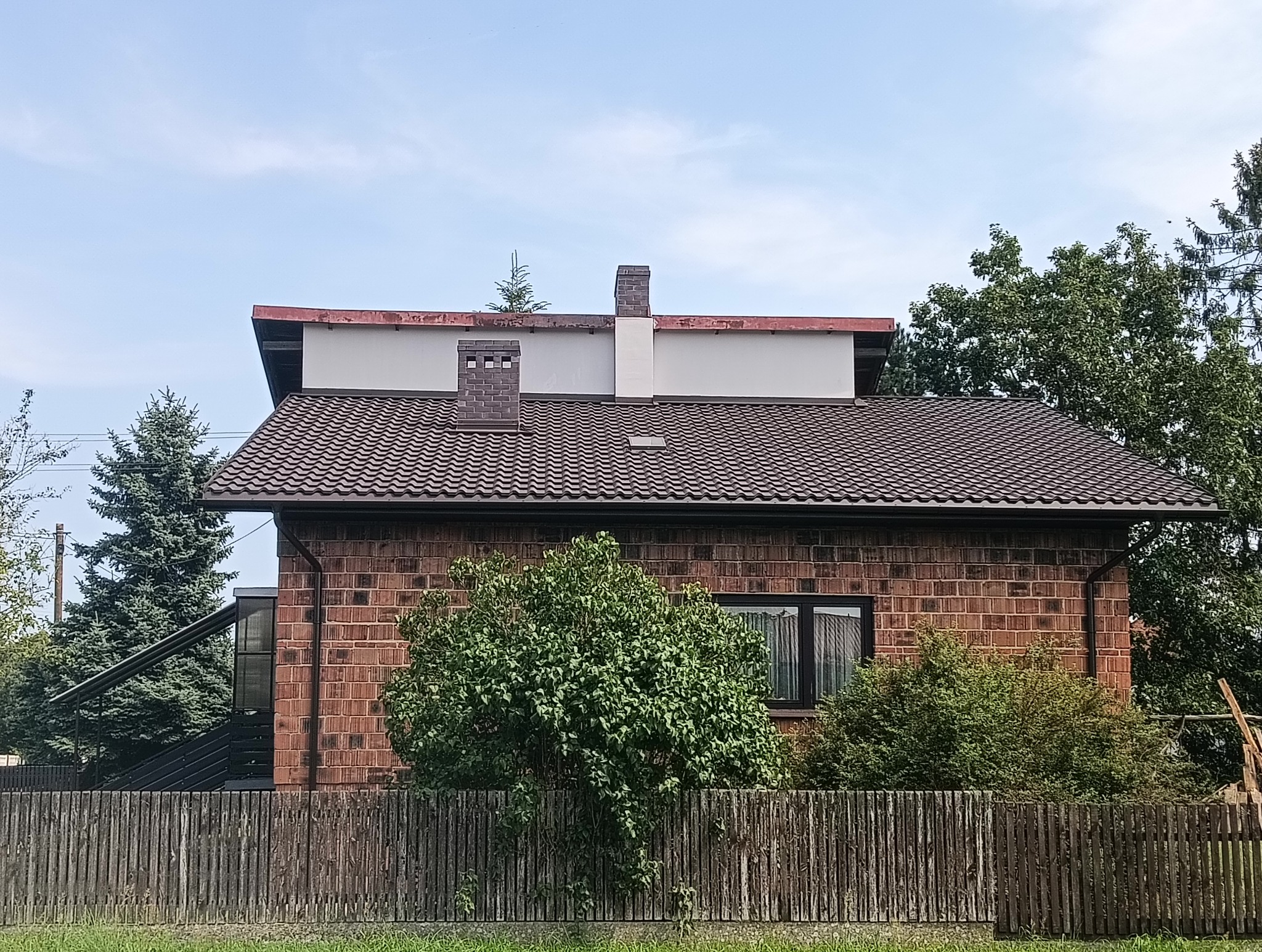 Dom z brązowym dachem z dachówki ceramicznej, ceglaną elewacją i nietypowymi kominami. W tle iglaste drzewa i błękitne niebo. Widok zza drewnianego płotu.