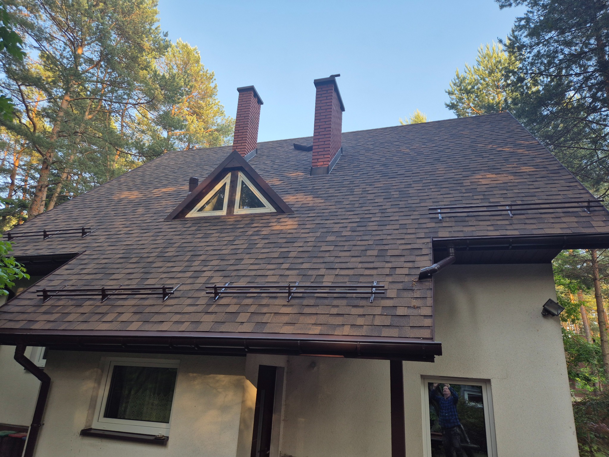 Brązowy dach z gontów bitumicznych, dwa kominy z czerwonej cegły, okno w kształcie trójkąta, rynny w kolorze dachu i drabinki przeciwśniegowe. Dom otoczony drzewami.