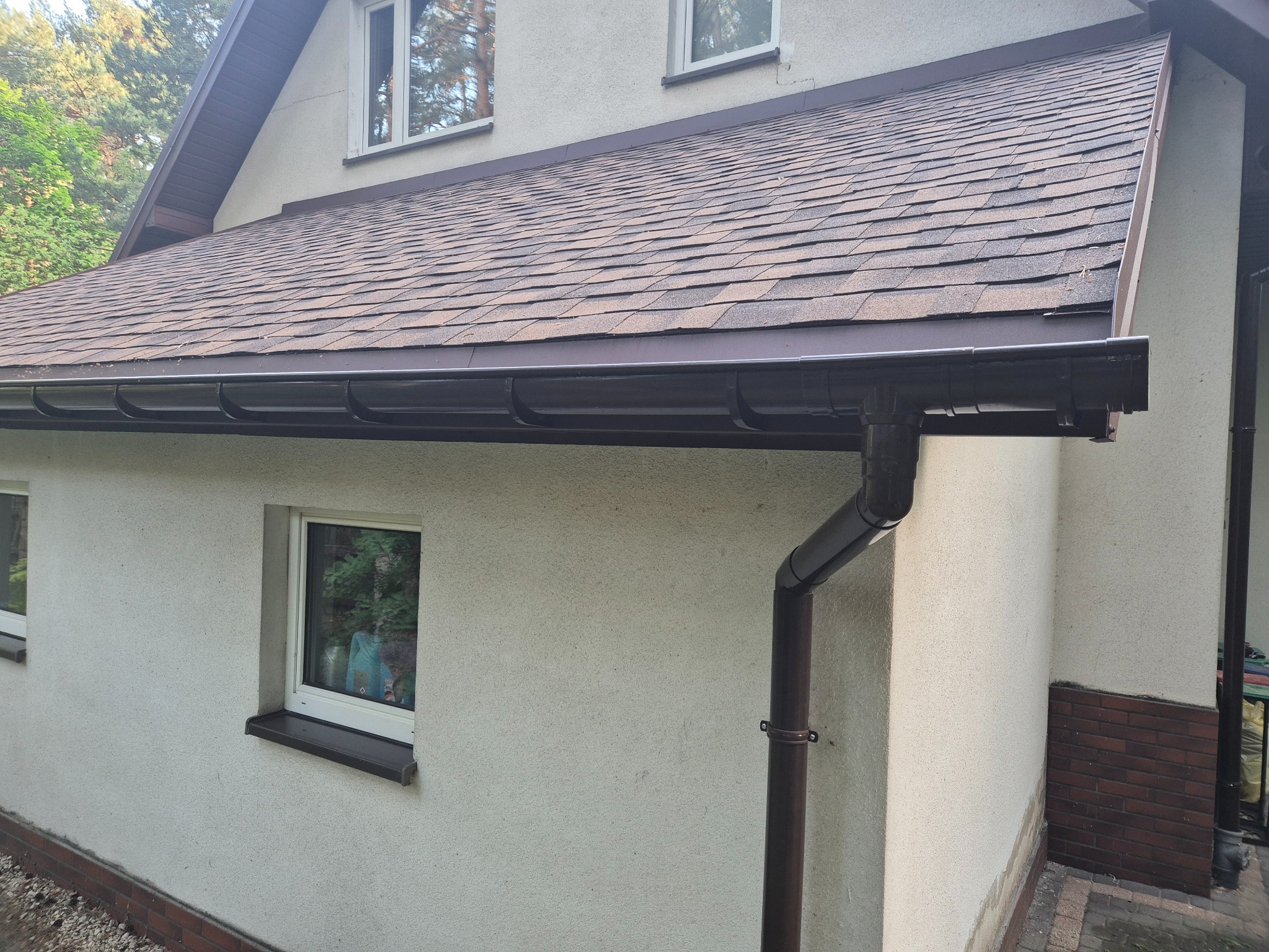 Dom z nowym, brązowym dachem pokrytym gontem bitumicznym i zamontowanym systemem rynnowym w tym samym kolorze. Widok z boku, z oknami i fragmentem elewacji.