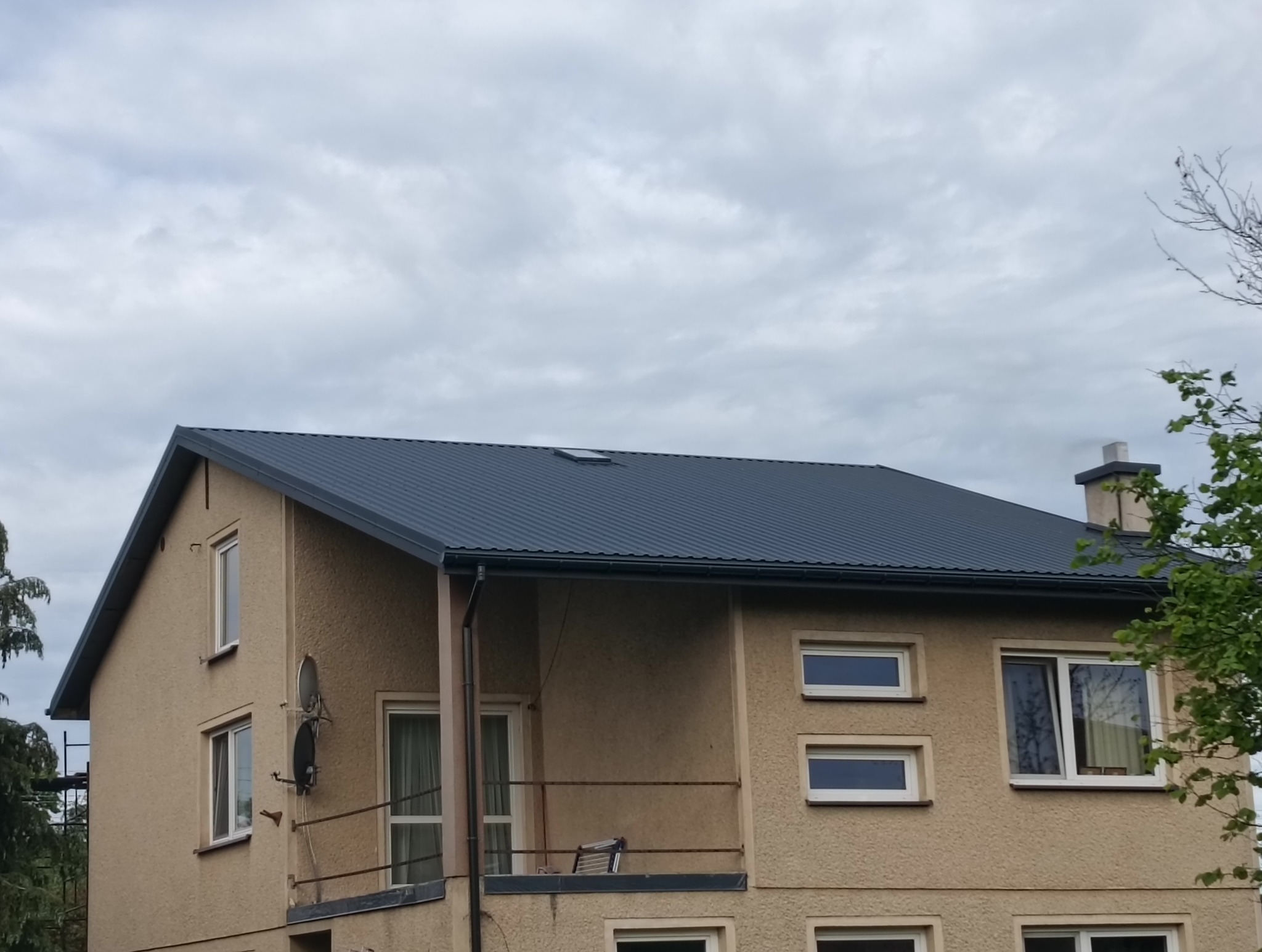 Dwupiętrowy dom z nowym, ciemnoszarym dachem z blachy trapezowej i oknem dachowym, na tle pochmurnego nieba. Widoczne rynny i komin.
