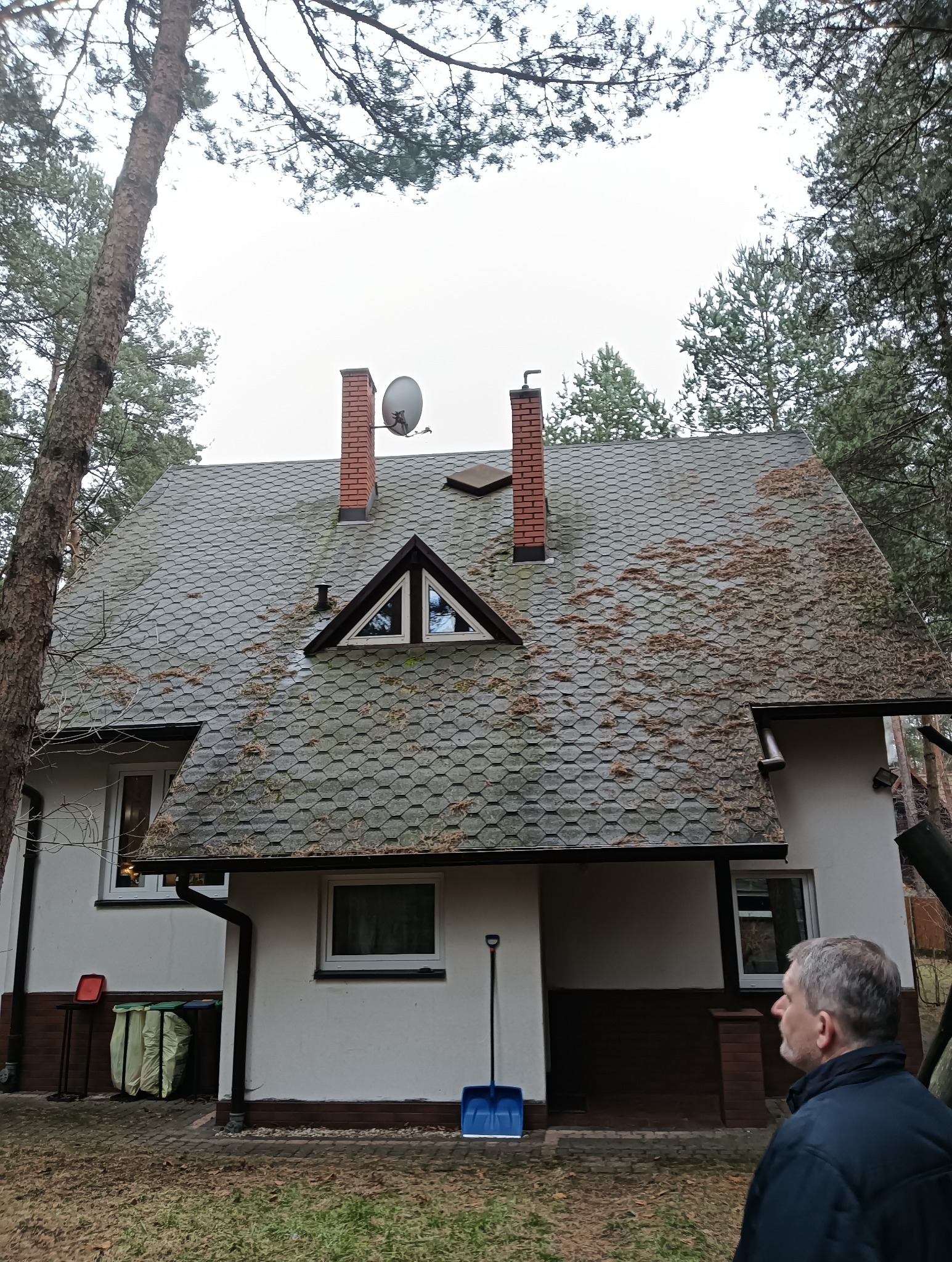 Dom z dachem pokrytym bitumiczną dachówką, częściowo porośniętym mchem. Widoczne kominy, antena satelitarna, rynny i drzewa. W tle pochmurne niebo. Na pierwszym planie mężczyzna.