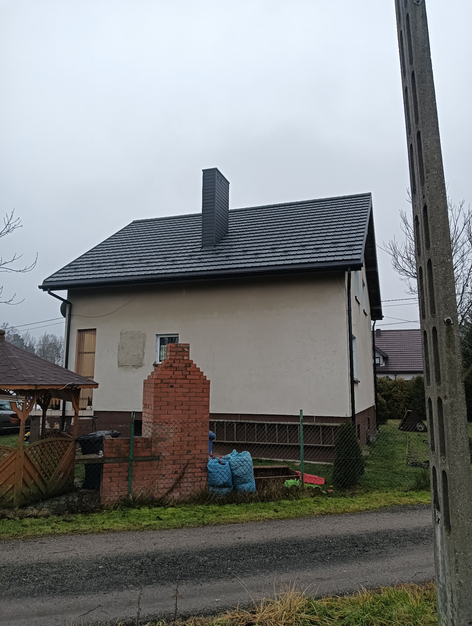 Dom jednorodzinny z nowym, ciemnym dachem i kominem. Widoczne obróbki blacharskie. Na pierwszym planie ceglana konstrukcja i worki. Pochmurne niebo.