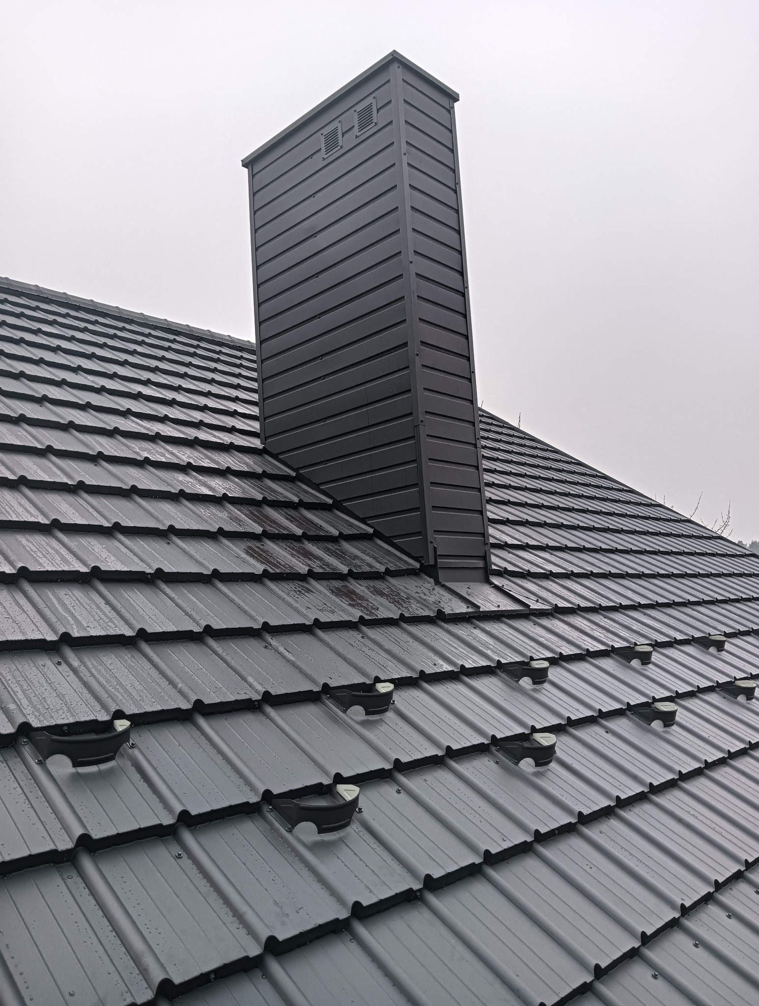 Szary, metalowy dach z kominem i zabezpieczeniami przeciwśniegowymi w pochmurny dzień. Widok z góry na nowo pokryty dach z blachy trapezowej.