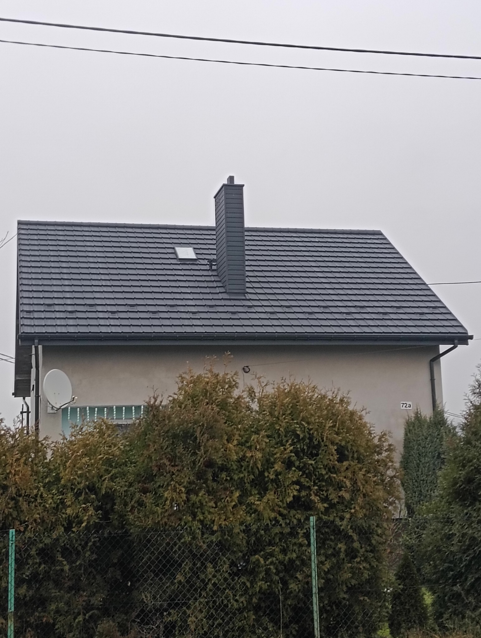 Dom z nowym, ciemnoszarym dachem z dachówki i kominem. Widoczne rynny, okno dachowe i antena satelitarna. Tło: pochmurne niebo, roślinność.