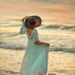 Our Little Stars - Kobieta w długiej, koronkowej sukience i kapeluszu na plaży o zachodzie słońca, trzymająca brzeg sukienki i kapelusz.