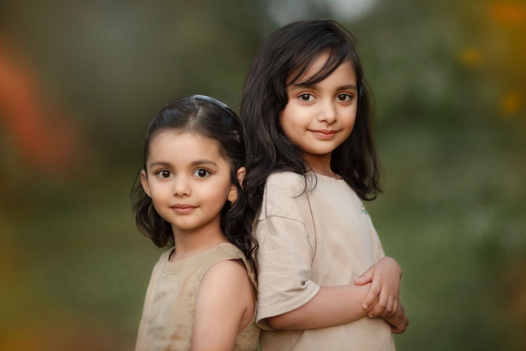 Portret dwóch uśmiechniętych dziewczynek opartych plecami do siebie na rozmytym, zielonym tle.