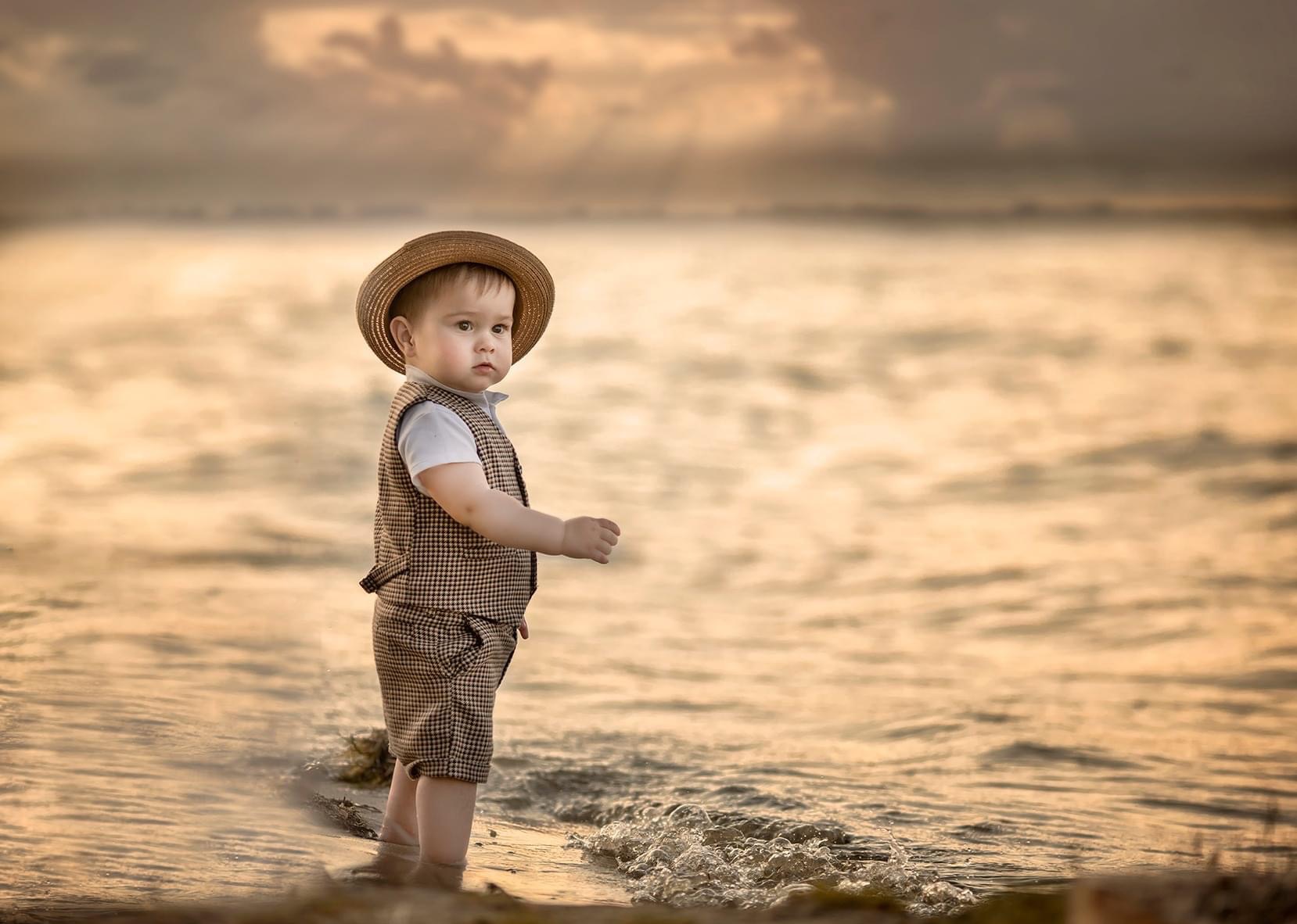 Mały chłopiec w stroju w kratę i słomkowym kapeluszu stoi w płytkiej wodzie na tle zachodzącego słońca.
