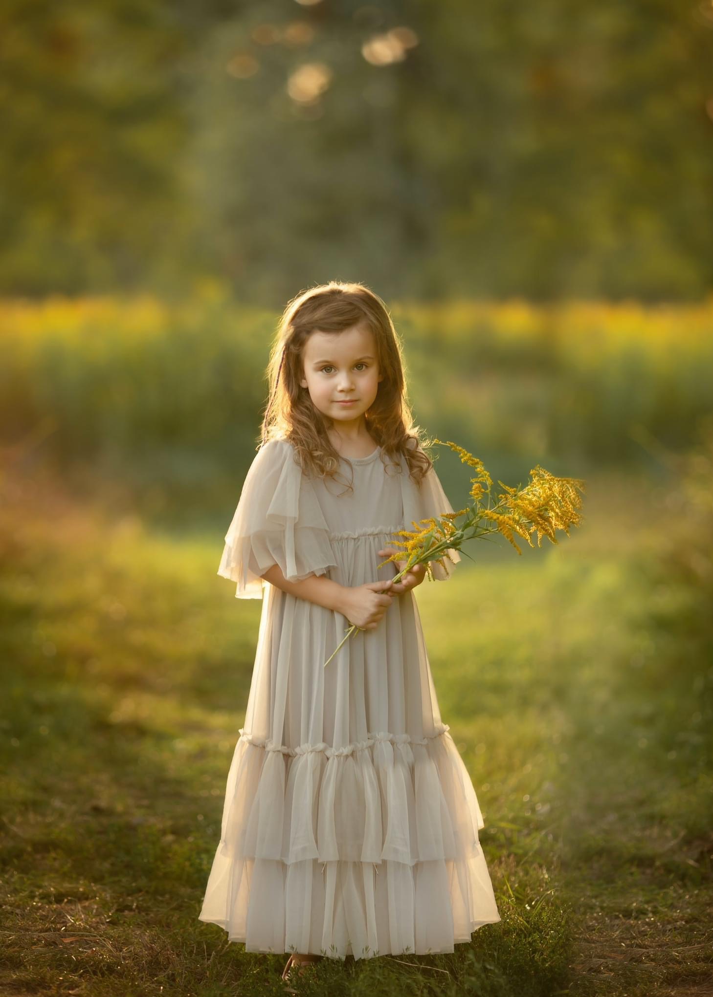 Portret dziewczynki w zwiewnej sukience trzymającej bukiet żółtych kwiatów na łące w złocistym świetle.