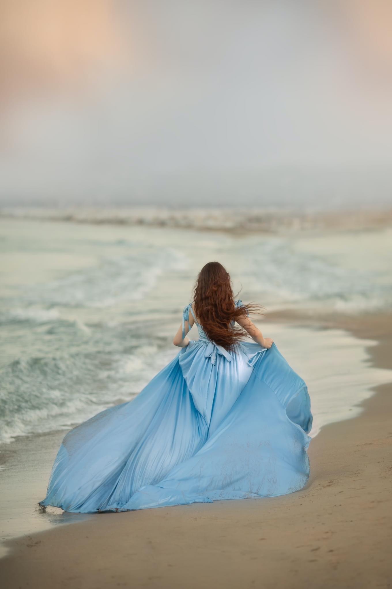 Kobieta w błękitnej, powiewnej sukni biegnie brzegiem morza, długie brązowe włosy falują na wietrze, widok z tyłu.
