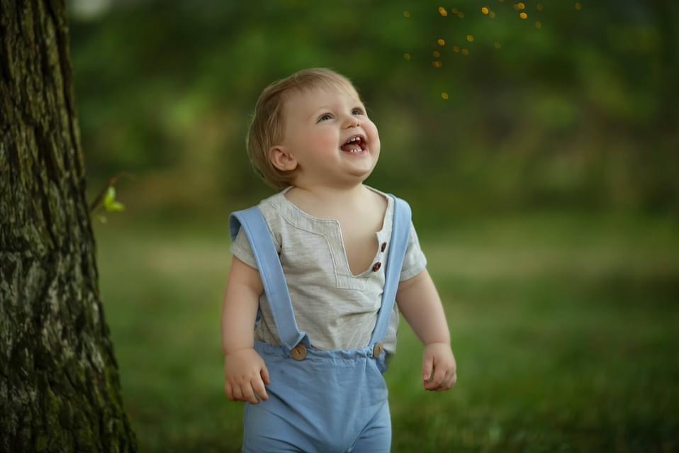 Uśmiechnięty chłopiec w błękitnych ogrodniczkach, z bluzką w kolorze ecru, stoi obok drzewa w parku w Warszawie, spoglądając w górę na rozproszone, złote punkciki światła.