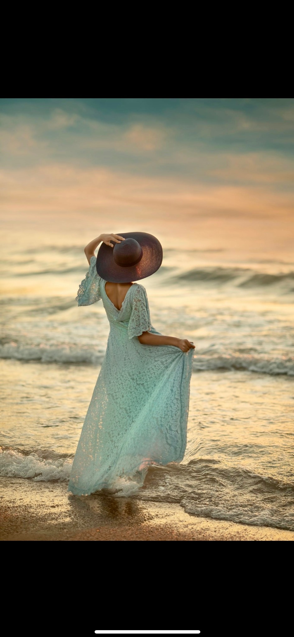 Kobieta w długiej, koronkowej sukience i kapeluszu na plaży o zachodzie słońca, trzymająca brzeg sukienki i kapelusz.