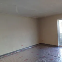 Świeżo otynkowane ściany w pomieszczeniu z folią grzewczą na podłodze, widoczne wystające kable z sufitu, przygotowane do dalszych prac wykończeniowych.