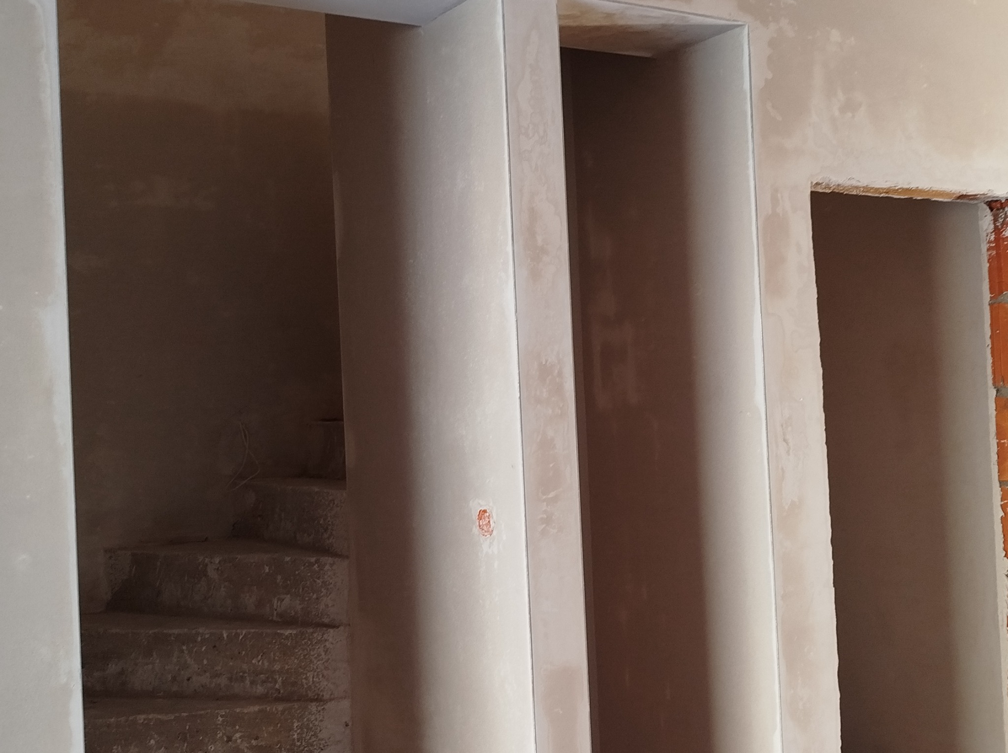 Surowe wnętrze w trakcie prac wykończeniowych: fragment schodów i przejścia w stanie deweloperskim, ściany pokryte świeżym, jasnym tynkiem, widoczne niedoskonałości i fragmenty cegieł.